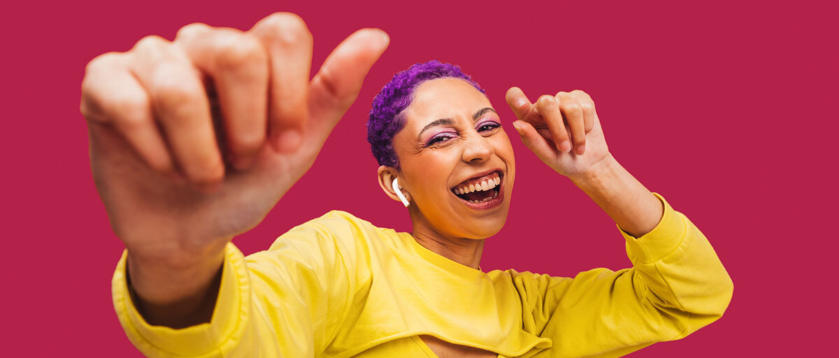 Eine Frau mit kurzen lila Haaren und leuchtend gelbem Shirt hört Musik über Kopfhörer und tanzt vor pinkem Hintergrund.