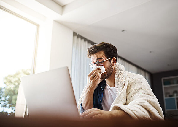 Erkälteter Mann sitzt mit einer Decke um den Körper, putzt sich die Nase und arbeitet mit seinem Laptop