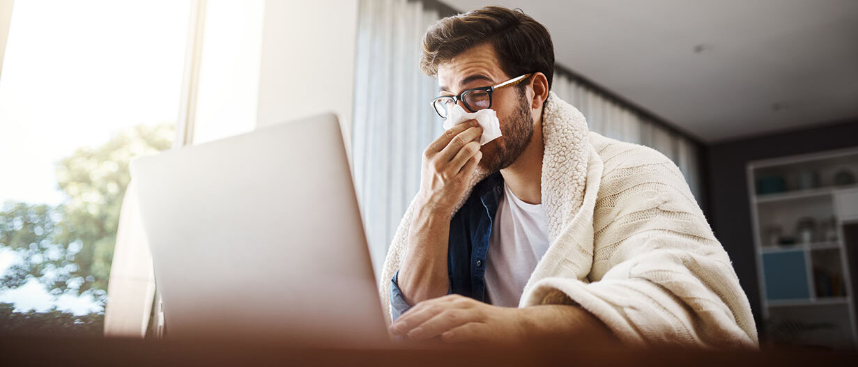 Präsentismus erkennen und vermeiden Erkälteter Mann sitzt mit einer Decke um den Körper, putzt sich die Nase und arbeitet mit seinem Laptop