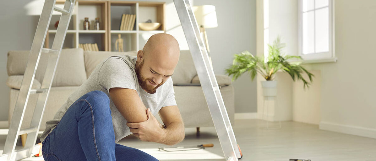 Ein Mann sitzt vor einer Leiter auf dem Wohnzimmerfußboden, neben ihm liegen Werkzeuge. Sein Gesicht ist schmerzverzerrt und er hält sich den Arm.