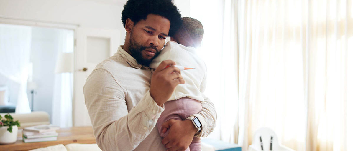 Ein Vater hält sein kleines Baby auf dem Arm und schaut besorgt auf ein Fieberthermometer.