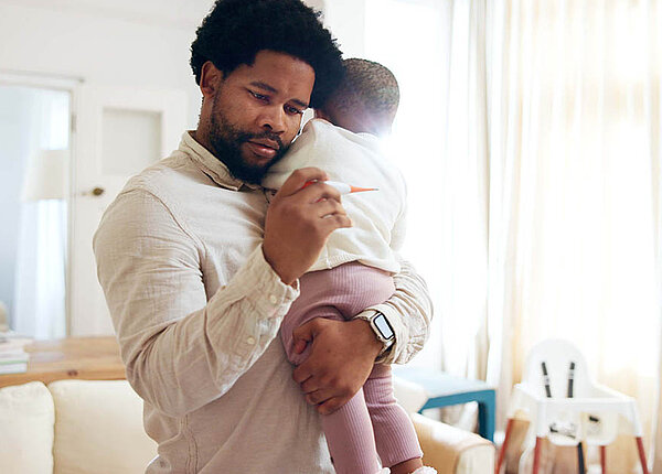 Ein Vater hält sein kleines Baby auf dem Arm und schaut besorgt auf ein Fieberthermometer.
