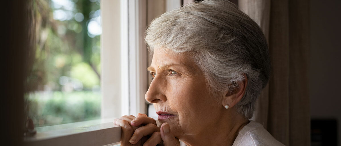 Ältere Frau steht am Fenster und schaut traurig
