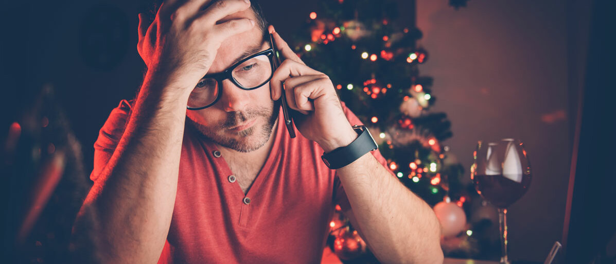 Ein Mann mit Brille sitzt, den Kopf in die Hände gestützt, über seinem Kalender und telefoniert. Neben ihm steht ein Glas Wein, das Licht ist schummrig, im Hintergrund leuchtet der Weihnachtsbaum.