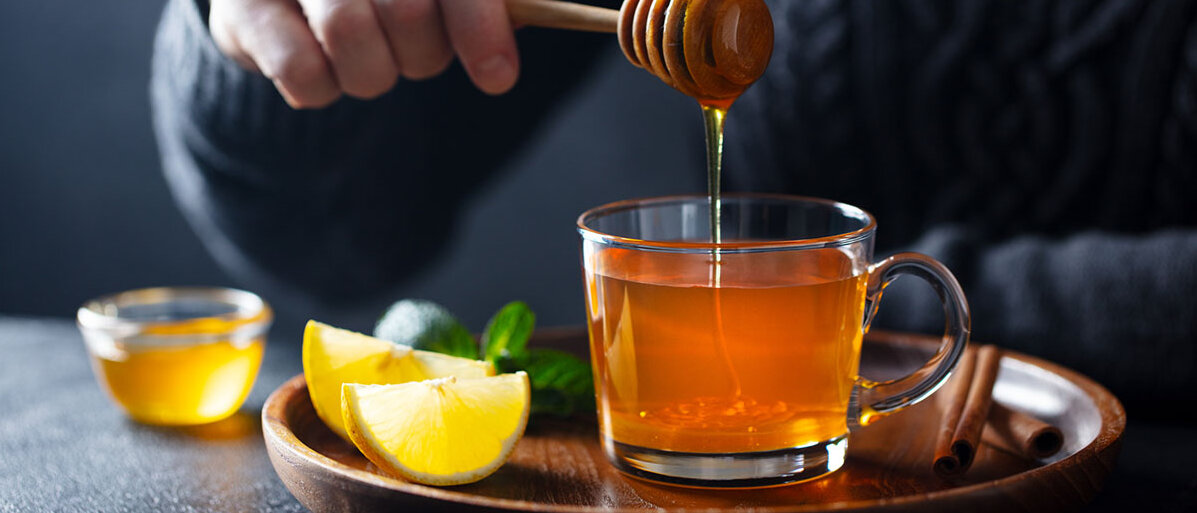 Honig als Hausmittel Mann mit Honiglöffel, der Honig in eine durchsichtige Teetasse gibt. Daneben Zitronenspalten.