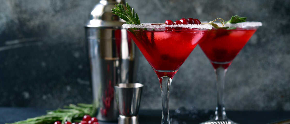 In zwei Martinigläsern befindet sich ein roter Drink, garniert mit Cranberries oder roten Johannisbeeren und Tannenzweigen. Die Beeren und Zweige liegen auch auf dem Tisch, im Hintergrund steht ein Cocktailshaker.
