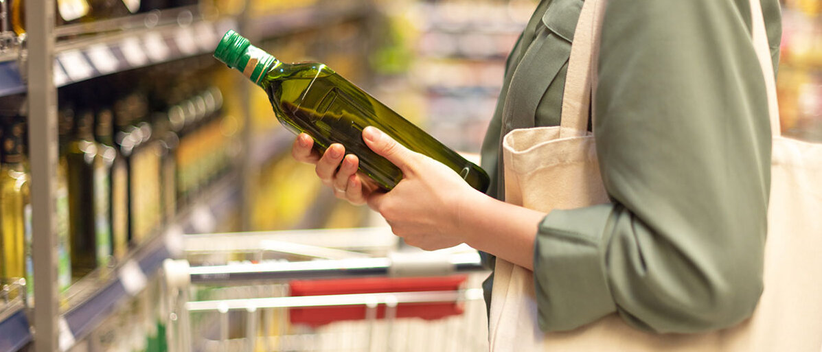 Eine Frau nimmt eine grüne Olivenölflasche aus einem Supermarktregal.