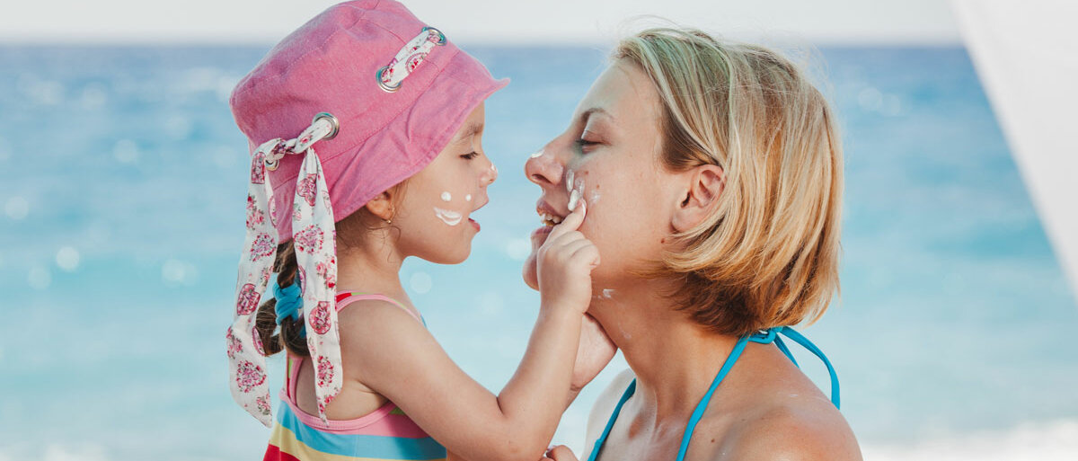 Eine Frau und ihr Kind sitzen am Strand und cremen sich gegenseitig das Gesicht mit Sonnencreme ein.