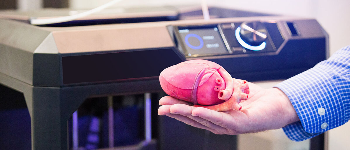 Mann hält ein menschliches Herz. Im Hintergrund sieht man einen 3D-Drucker.