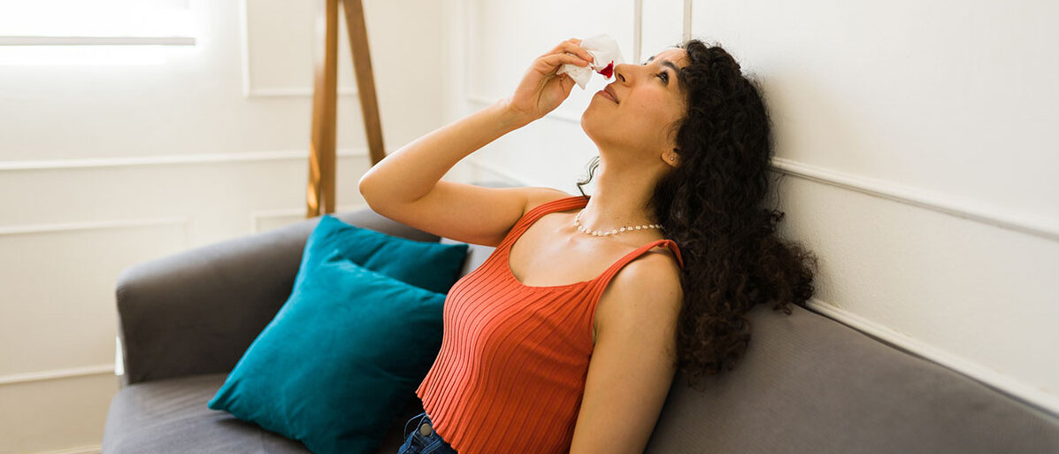 Nasenbluten Frau mit Nasenbluten sitzt auf einem Sofa und legt den Kopf in den Nacken
