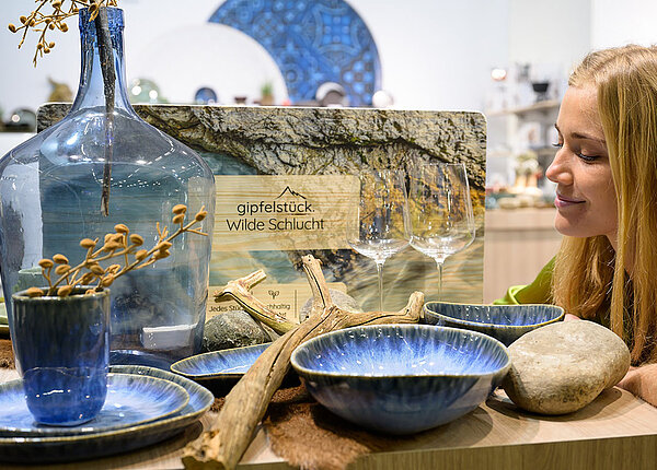 Auf einem Messe-Ausstellungstisch steht blaues Geschirr im nordisch-unperfekten Stil und eine große, blaue Vase. Eine Frau beugt sich darüber und begutachtet die Ausstellungsstücke.