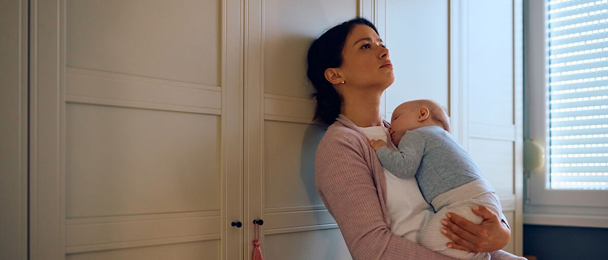 Traurig wirkende Mutter hat ihr schlafendes Baby auf dem Arm und ist gegen einen Schrank gelehnt