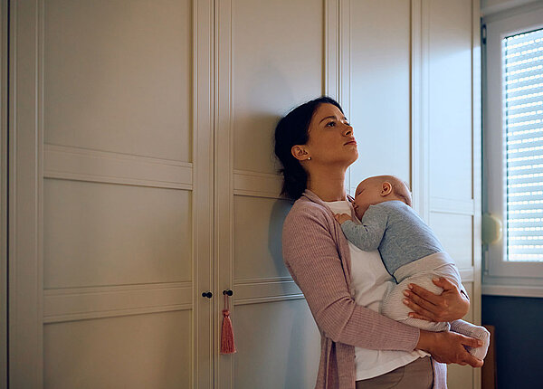 Wochenbettdepression oder Babyblues? So erkennen Sie den Unterschied Traurig wirkende Mutter hat ihr schlafendes Baby auf dem Arm und ist gegen einen Schrank gelehnt