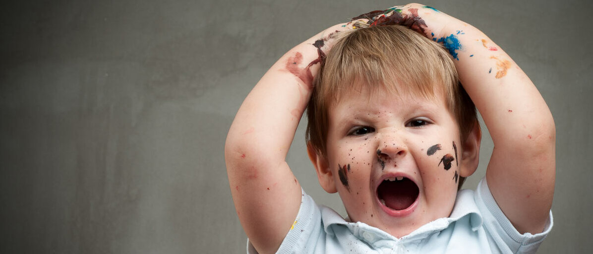 Ein Junge im Kindergartenalter schlägt die Hände über dem Kopf zusammen und verzieht aufgebracht das Gesicht. Auf seinem Gesicht, den Armen und seinem Shirt sind viele Flecken von Malfarbe.