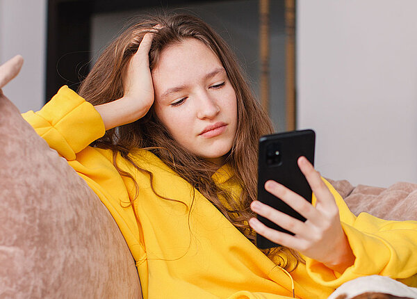 Eine Jugendliche sitzt auf dem Sofa. Sie hat den Arm auf der Lehne abgestützt und ihren Kopf auf die Hand gestützt. Dabei blickt sie missmutig in ihr Smartphone.