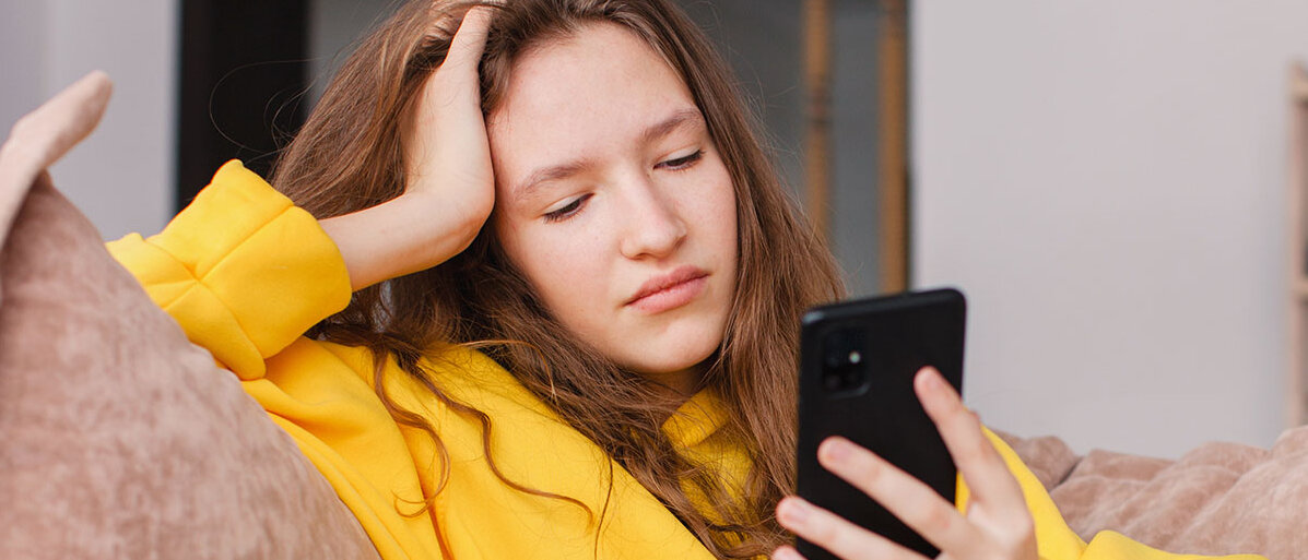 Eine Jugendliche sitzt auf dem Sofa. Sie hat den Arm auf der Lehne abgestützt und ihren Kopf auf die Hand gestützt. Dabei blickt sie missmutig in ihr Smartphone.