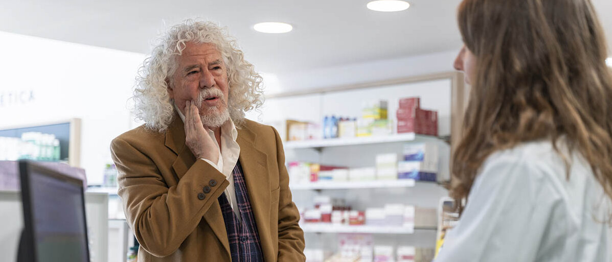 Ein Kunde steht vor dem HV-Tisch und wird beraten. Er hält die Hand an die Wange, weil er Zahnschmerzen hat.