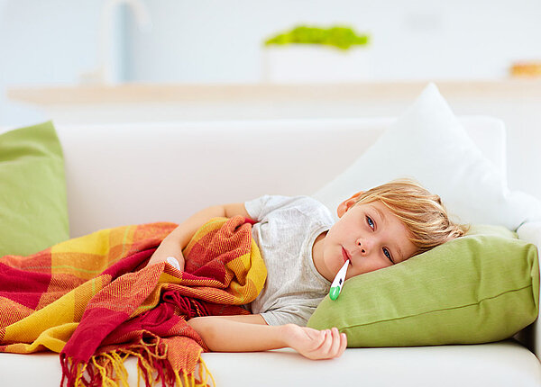 Gefährliches Fieber bei Kindern erkennen Kranker kleiner Junge liegt mit Fieberthermometer im Mund auf der Couch.