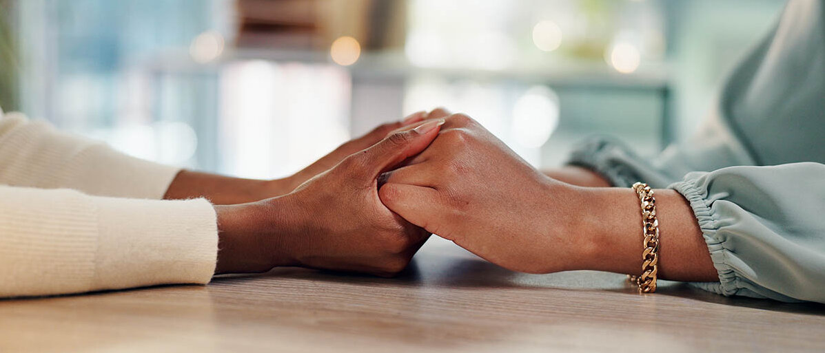 Empathie-Training: Wie Mitgefühl zu lernen das Beratungsgespräch und die Kommunikation in der Apotheke verbessern kann Zwei Frauen halten sich über einen Tisch hinweg an den Händen.