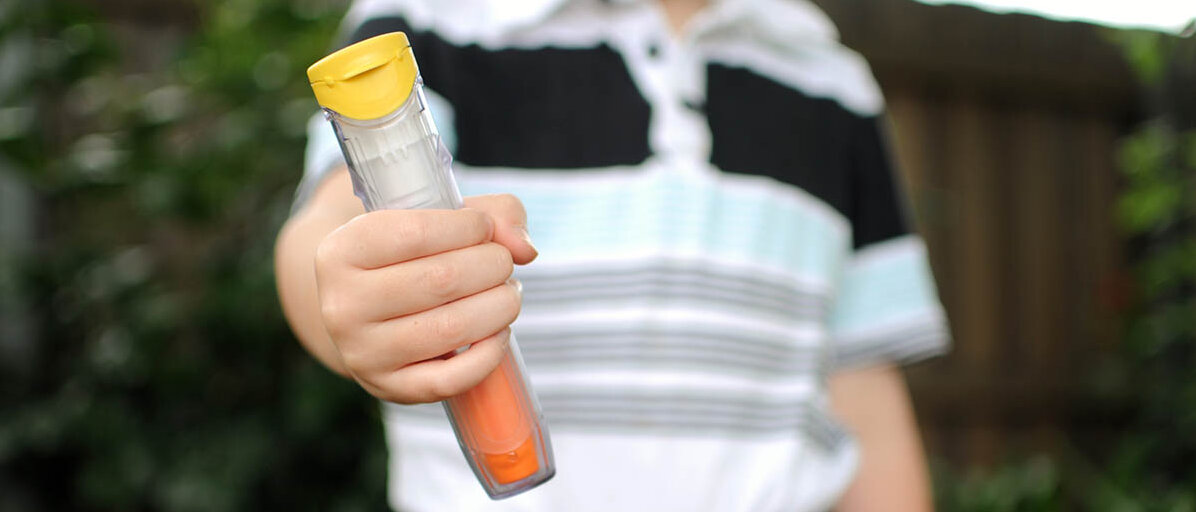 Ein Junge hält den Betrachtenden einen gelb-orangen Epipen entgegen. Einen weiteren Pen hat er in seiner Bauchtasche.