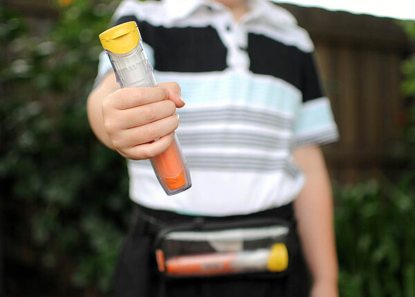 Ein Junge hält den Betrachtenden einen gelb-orangen Epipen entgegen. Einen weiteren Pen hat er in seiner Bauchtasche.