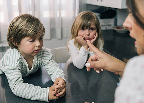 Verbale Gewalt an Kindern ist Misshandlung. Eine Frau spricht mit erhobenem Finger mit zwei Kindern im Kindergartenalter. Beide sehen zerknirscht aus.