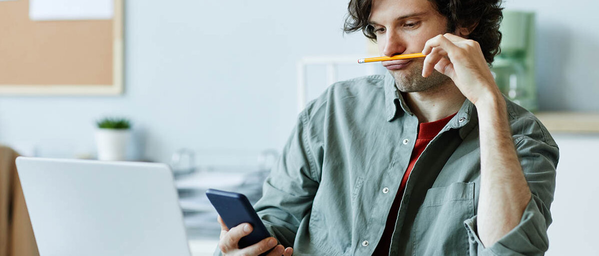 Ein Mann in den Dreißigern sitzt vor dem aufgeklappten Laptop, schaut auf sein Handy und spielt dabei mit einem Bleistift herum.