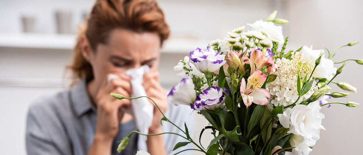Frau sitzt hinter buntem Blumenstrauß und muss sich die Nase schnäuzen