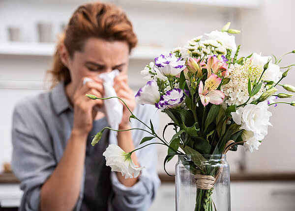 Frau sitzt hinter buntem Blumenstrauß und muss sich die Nase schnäuzen