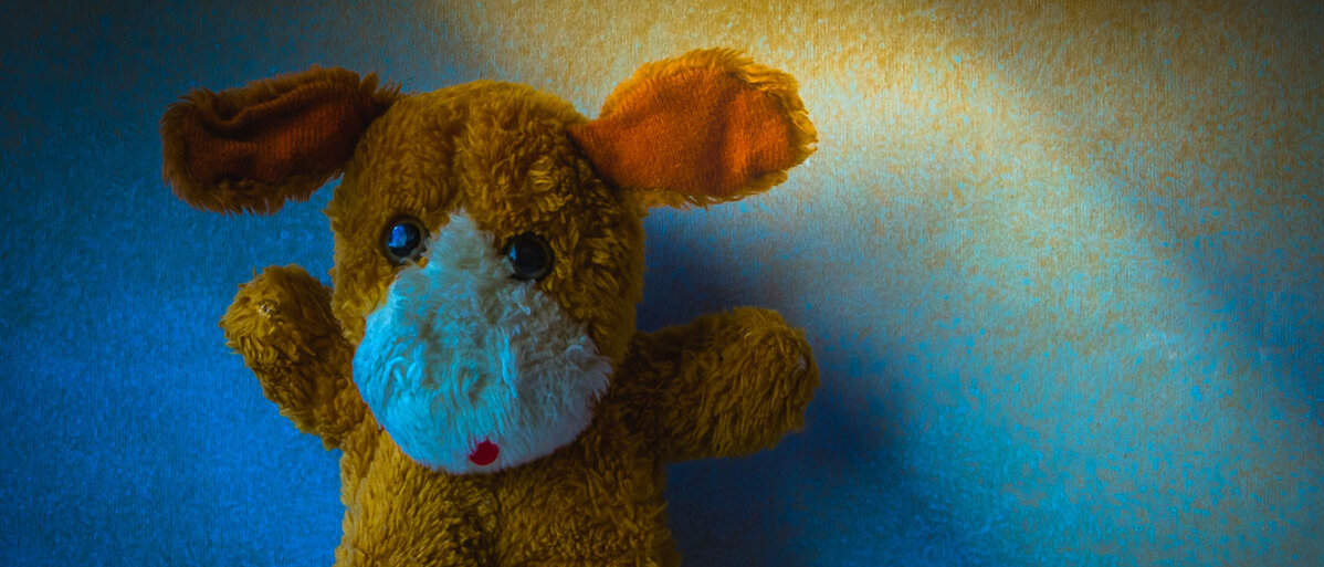 Ein Kuscheltier steht allein in einem dunklen Raum, nur erhellt von einem Lichtstrahl. Es hat große runde Augen und hebt die Arme nach oben.