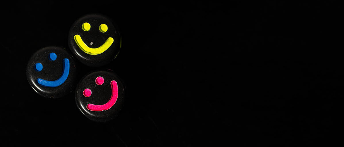 Drei schwarze Pillen mit Smileys in pink, gelb und blau vor einem schwarzen Hintergrund