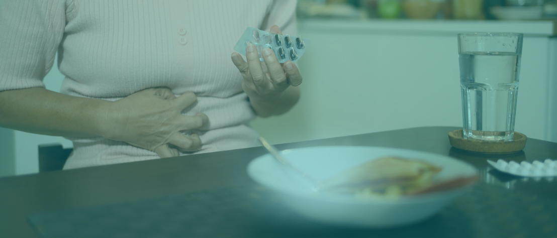 Eine Frau hat einen halb aufgegessenen Teller vor sich stehen, greift sich verkrampft an den Bauch und hält einen angebrochenen Blister mit Kapseln in der Hand. Der Fokus liegt auf dem Essen.