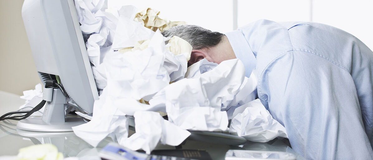 Mann liegt mit seinem Kopf auf seinem Laptop in einem Meer aus zerknülltem Papier