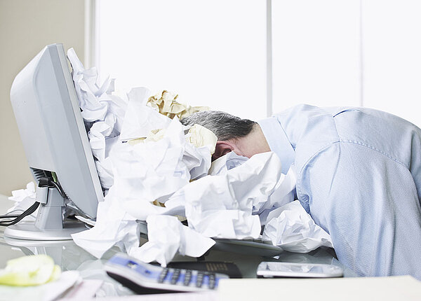 Mann liegt mit seinem Kopf auf seinem Laptop in einem Meer aus zerknülltem Papier