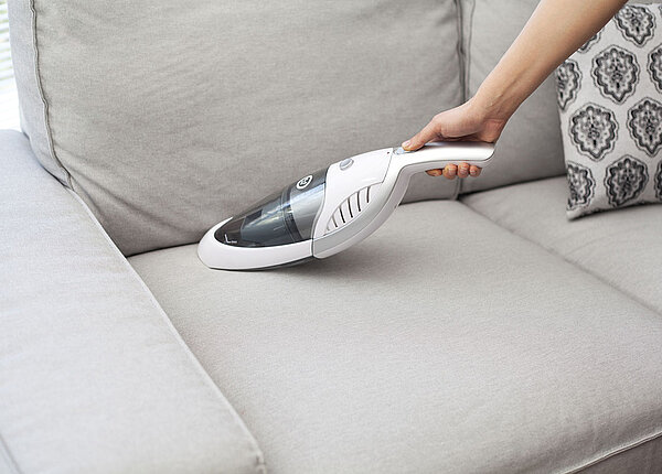 Frau reinigt graues Sofa mit Handstaubsauger