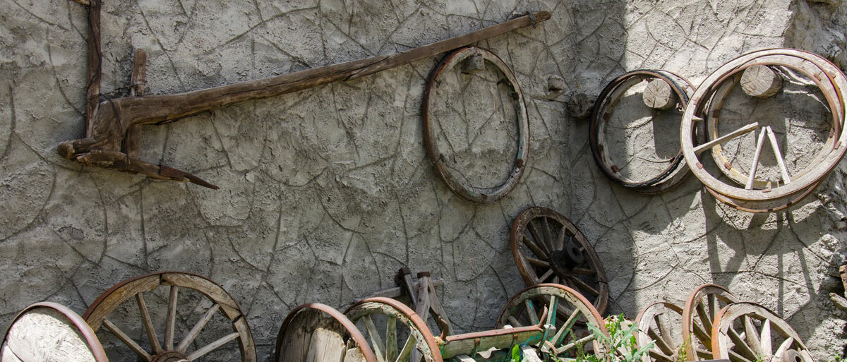 An einer alten Mauer lehnen und hängen hölzerne historische Arbeitsgeräte für den Ackerbau, zum Beispiel mehrere Räder und ein Pflug.
