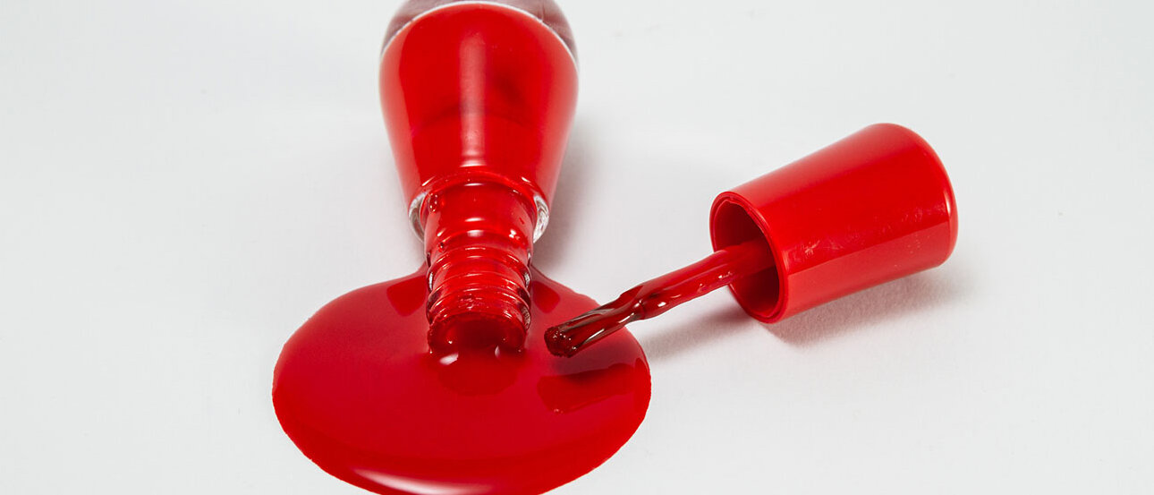 Nagellack richtig entsorgen Roter Nagellack aus einer Glasflasche, der ausgelaufen ist. Nebendran liegt der Pinsel.