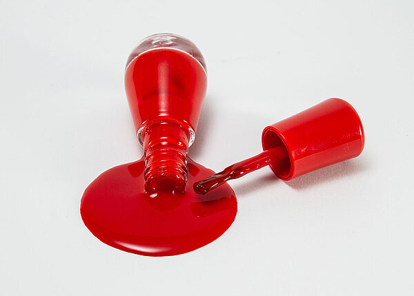 Roter Nagellack aus einer Glasflasche, der ausgelaufen ist. Nebendran liegt der Pinsel.