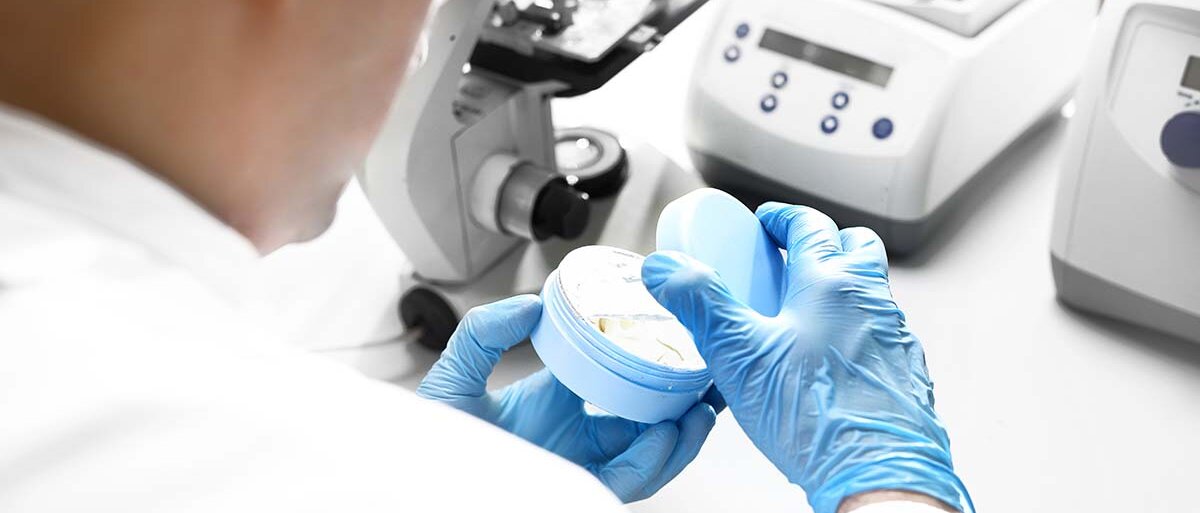 Ein Mann in weißem Kittel steht vor einem Labortisch mit Mikroskop und weiteren technischen Geräten. Er öffnet einen Cremetiegel und begutachtet den Inhalt.