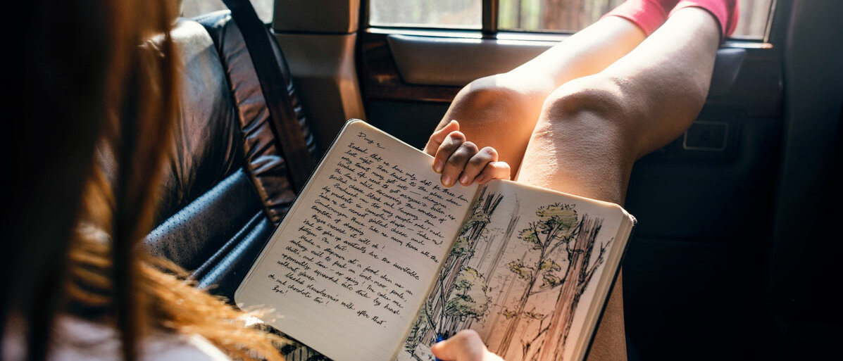 Eine junge Frau sitzt im Auto, lässt die Beine aus dem Fenster baumeln und schreibt und zeichnet in ein Notizbuch.