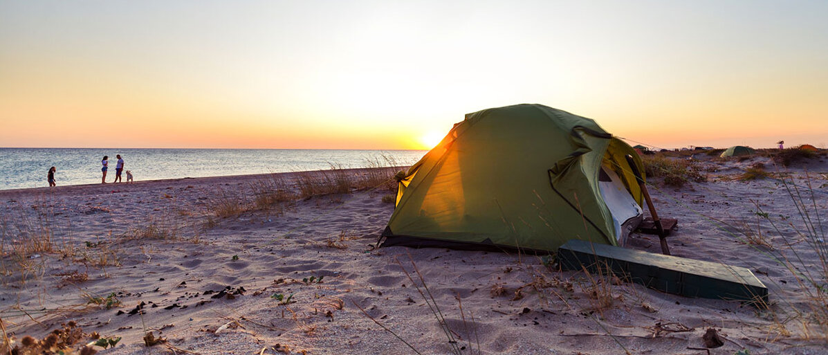 Sonnenuntergang am Strand: Im Sand steht ein Zelt, an der Wasserkante spazieren drei Menschen und ein Hund.