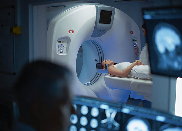 Eine Ärztin oder MT schiebt eine Patientin ins MRT. Vom Kontrollraum aus schaut ein Arzt zu.