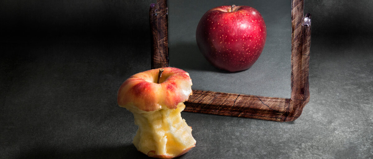 Ein bereits abgegessener Apfel steht vor dem Spiegel. Das Spiegelbild zeigt einen vollen, runden Apfel.