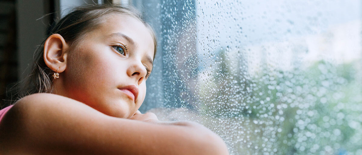 Mädchen schaut traurig aus dem Fenster. Am Fenster sind Regentropfen
