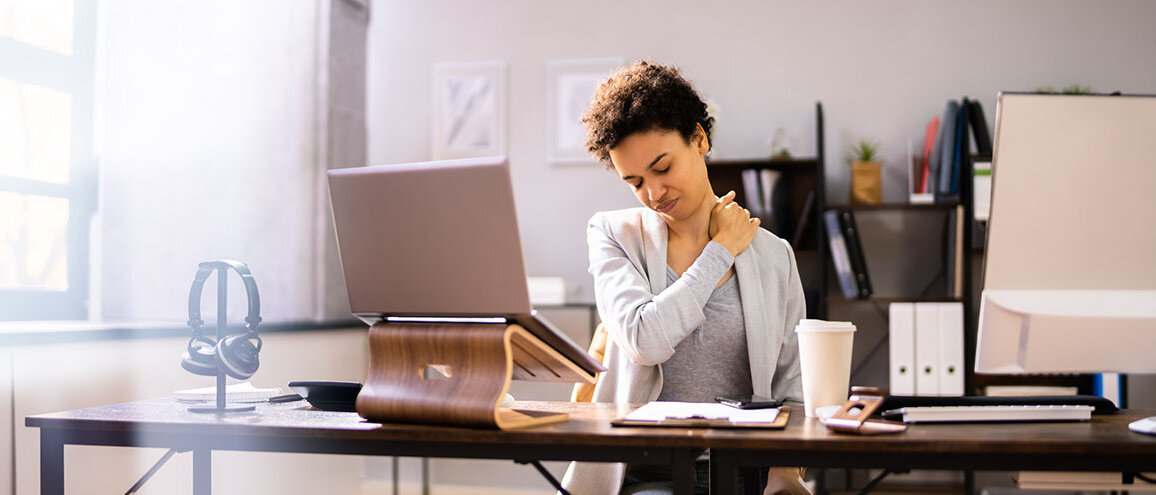 Frau sitzt am Schreibtisch vorm PC und hält sich den Schulter-Nackenbereich