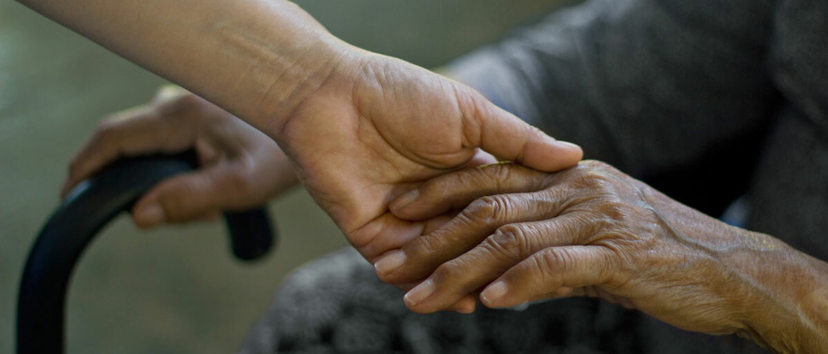 Eine junge Person hält die Hand einer alten, sitzenden Person. Die ältere Person hält in der anderen Hand einen Gehstock.