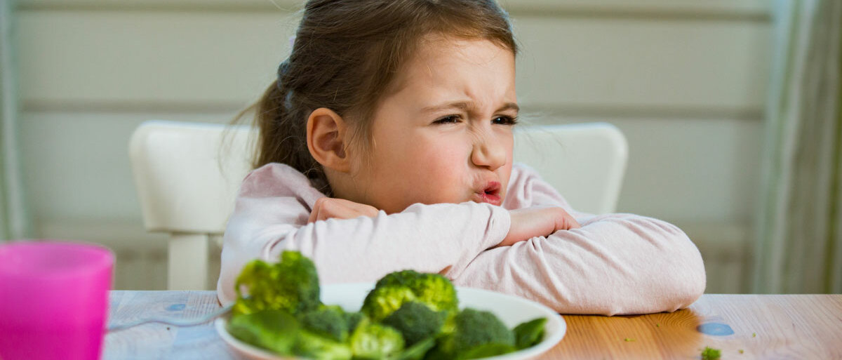 Ein Mädchen sitzt vor einem Teller mit Brokkoli, verschränkt die Arme und verzieht das Gesicht.