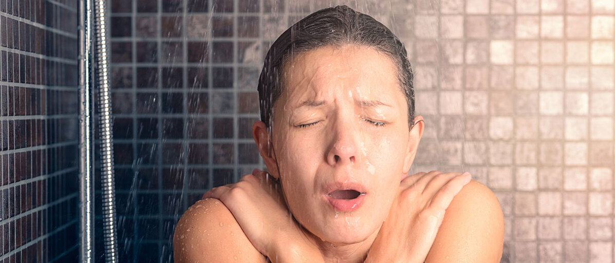 Eine Frau nimmt eine kalte Dusche: Sie kneift die Augen zusammen, hat den Mund zu einem O geformt, den Kopf eingezoge und verkreuzt die Arme vor dem Oberkörper.