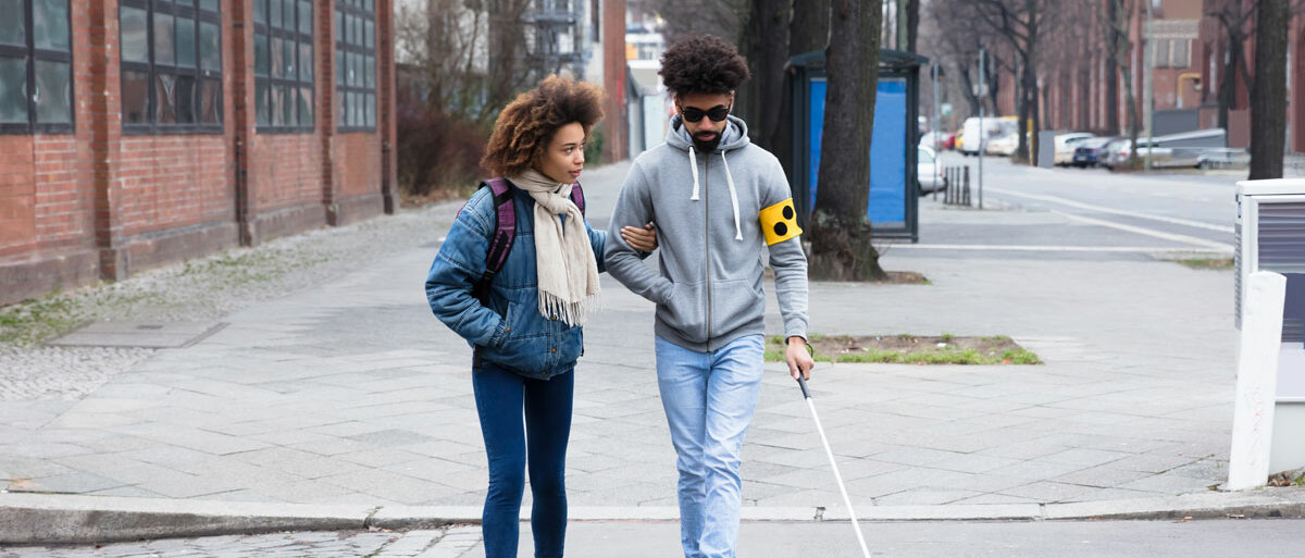 Ein junger Mann trägt eine gelbe Armbinde mit drei schwarzen Punkten und eine dunkle Sonnenbrille. Mit Hilfe eines Blindenstocks und am Arm einer jungen Frau überquert er eine Straße.