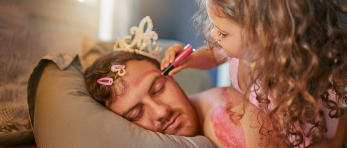 Ein Mann liegt tagsüber im Bett und schläft. Seine Tochter hat ihm eine Krone aufgesetzt und bemalt ihn im Gesicht mit pinkem Filzstift.